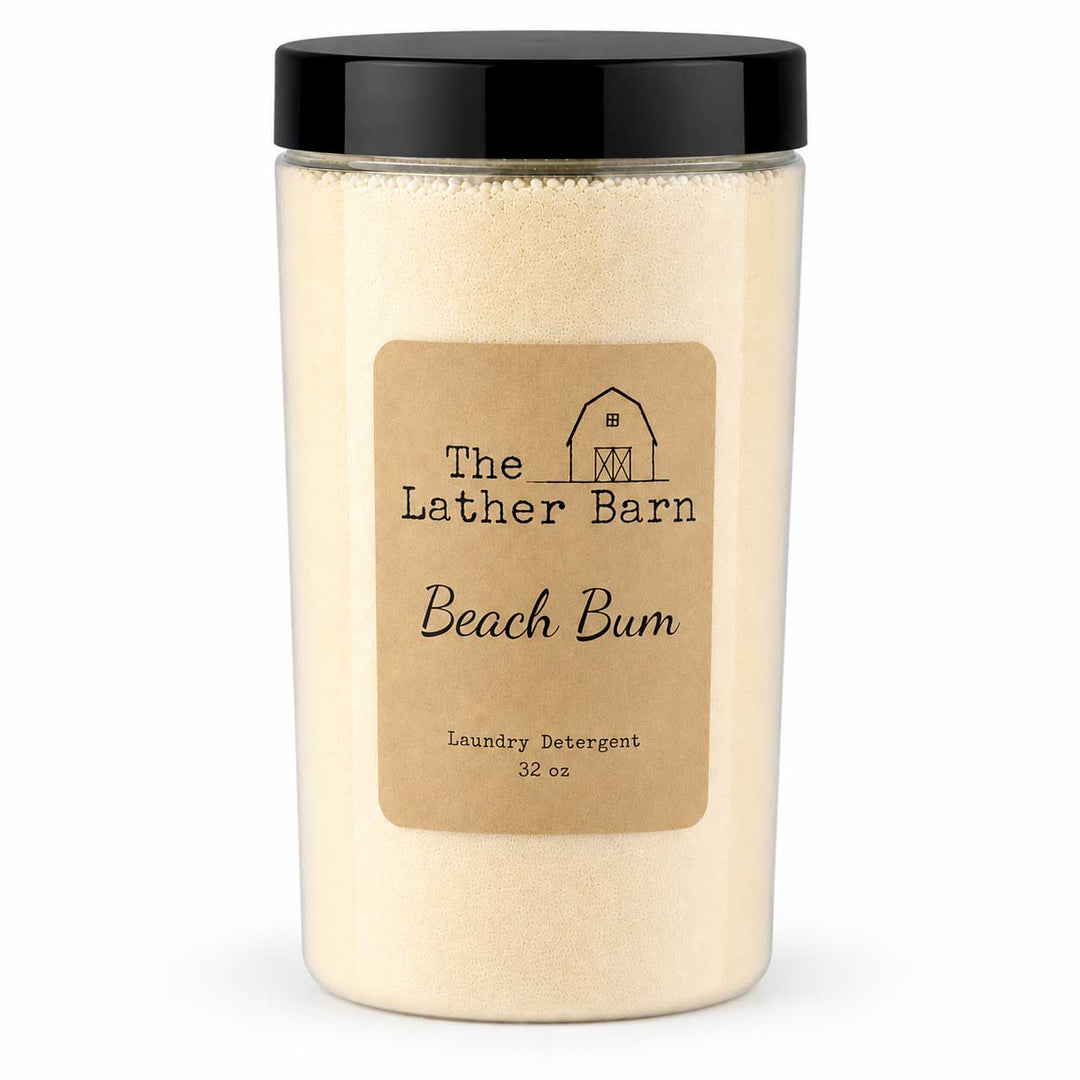 A 32 oz jar of The Lather Barn Beach Bum laundry detergent, featuring a clear container filled with a light beige powder, a tan paper label, and a black screw-on lid, set against a plain white background.