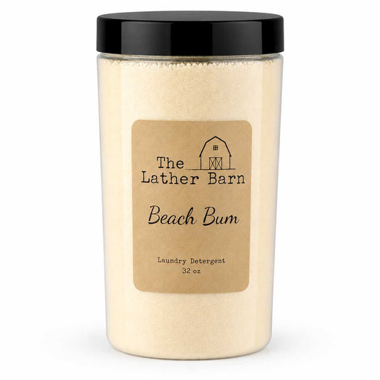 A 32 oz jar of The Lather Barn Beach Bum laundry detergent, featuring a clear container filled with a light beige powder, a tan paper label, and a black screw-on lid, set against a plain white background.