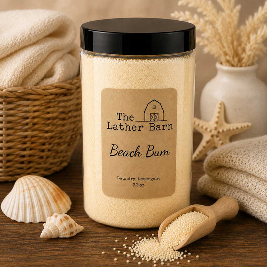 A jar of The Lather Barn Beach Bum laundry detergent sits on a wooden table surrounded by beach-themed props including seashells, a wooden scoop, folded towels, and dried grass in a vase.