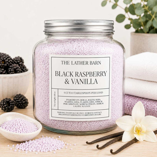 A large clear glass jar filled with pale lavender bath beads sits on a tabletop in a styled setting. The jar has a silver metal lid and a white label reading 'THE LATHER BARN BLACK RASPBERRY & VANILLA.