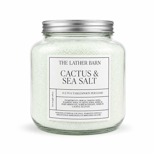 A clear glass jar filled with pale sea salt crystals and topped with a silver metal lid, labeled 'THE LATHER BARN CACTUS & SEA SALT' on a white background.