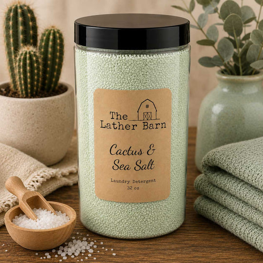 A jar of The Lather Barn Cactus & Sea Salt laundry detergent sits on a wooden table surrounded by a cactus, folded towels, and a small wooden scoop of salt.