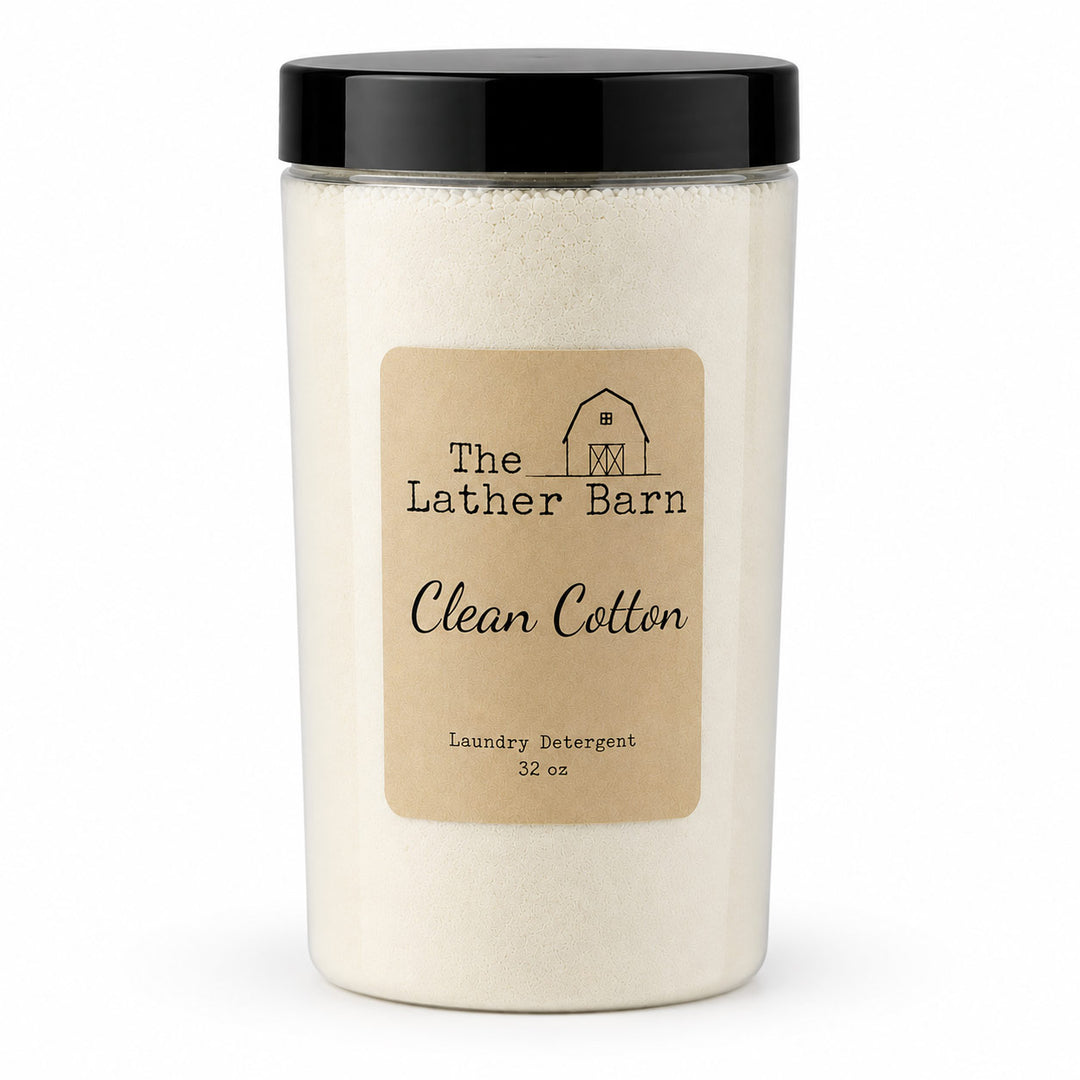 A 32 oz jar of The Lather Barn Clean Cotton laundry detergent. The product is shown in a clear plastic jar filled with off-white powder, featuring a black screw-on lid and a tan paper label with black text and a small barn illustration.