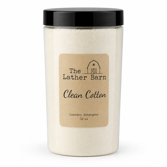 A 32 oz jar of The Lather Barn Clean Cotton laundry detergent. The product is shown in a clear plastic jar filled with off-white powder, featuring a black screw-on lid and a tan paper label with black text and a small barn illustration.