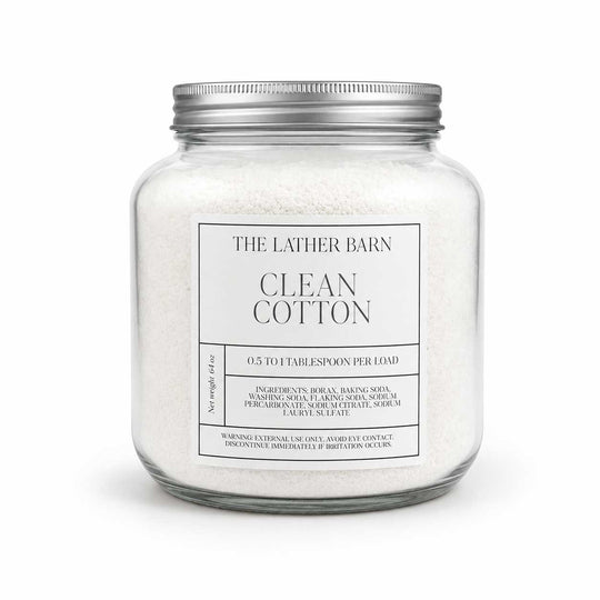 A glass jar candle filled with white cotton bath salts, featuring a silver metal lid and a white label that reads 'THE LATHER BARN CLEAN COTTON'.