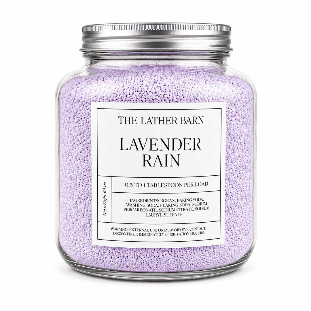 A clear glass jar candle filled with light lavender-colored granules, featuring a silver metal lid and a white label that reads 'THE LATHER BARN LAVENDER RAIN'.