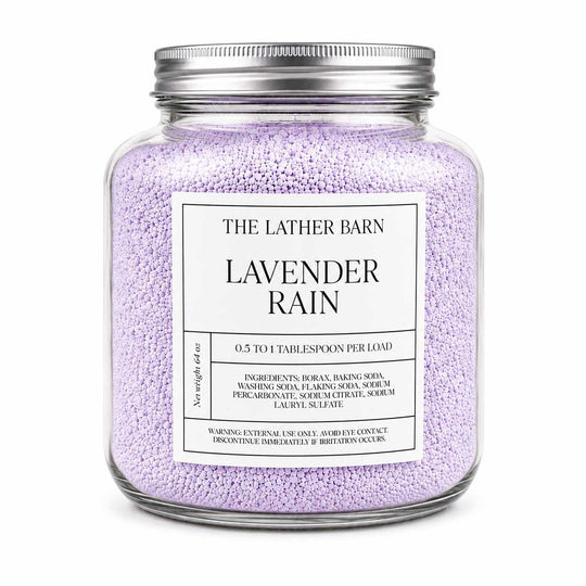 A clear glass jar candle filled with light lavender-colored granules, featuring a silver metal lid and a white label that reads 'THE LATHER BARN LAVENDER RAIN'.