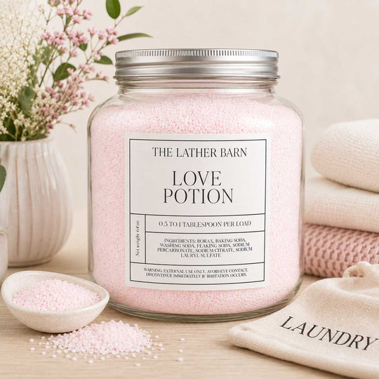 A glass jar candle filled with pale pink bath-like granules, topped with a silver metal lid, and labeled 'THE LATHER BARN LOVE POTION' on a white background.