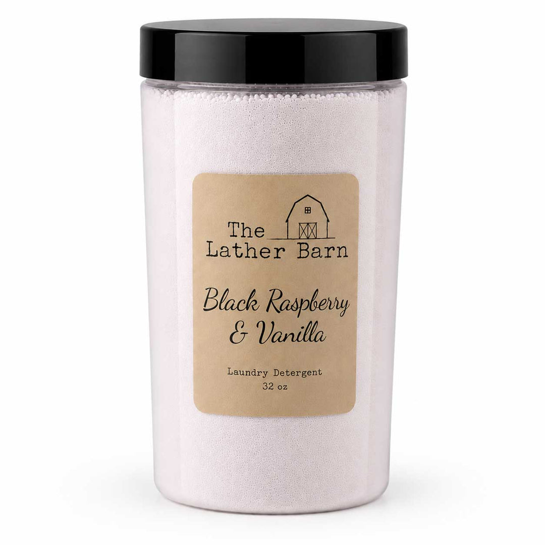 A jar of laundry detergent from The Lather Barn, featuring a black lid and a beige label with a barn logo. The product is labeled 'Black Raspberry & Vanilla' and contains light pinkish-beige detergent crystals.