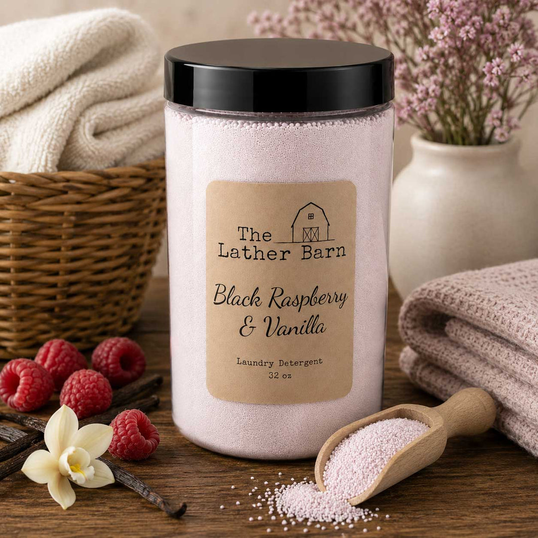 A jar of The Lather Barn Black Raspberry & Vanilla laundry detergent sits on a wooden table surrounded by bathhouse props, including folded towels, raspberries, vanilla beans, a flower, and a scoop of the pink powder.