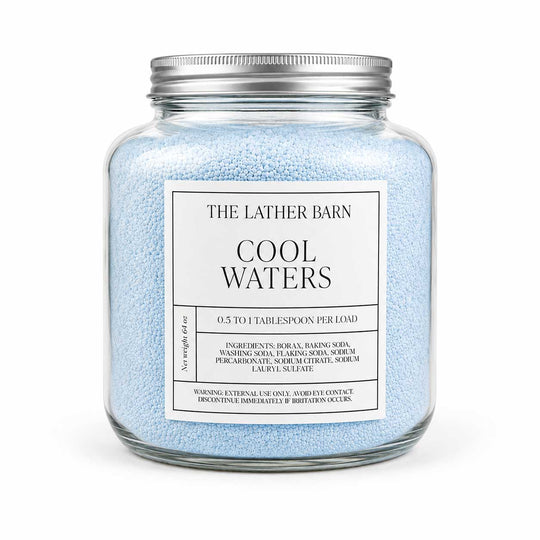 A glass jar filled with light blue bath salts, featuring a white label that reads 'THE LATHER BARN COOL WATERS' and a silver metal lid, set against a plain white background.