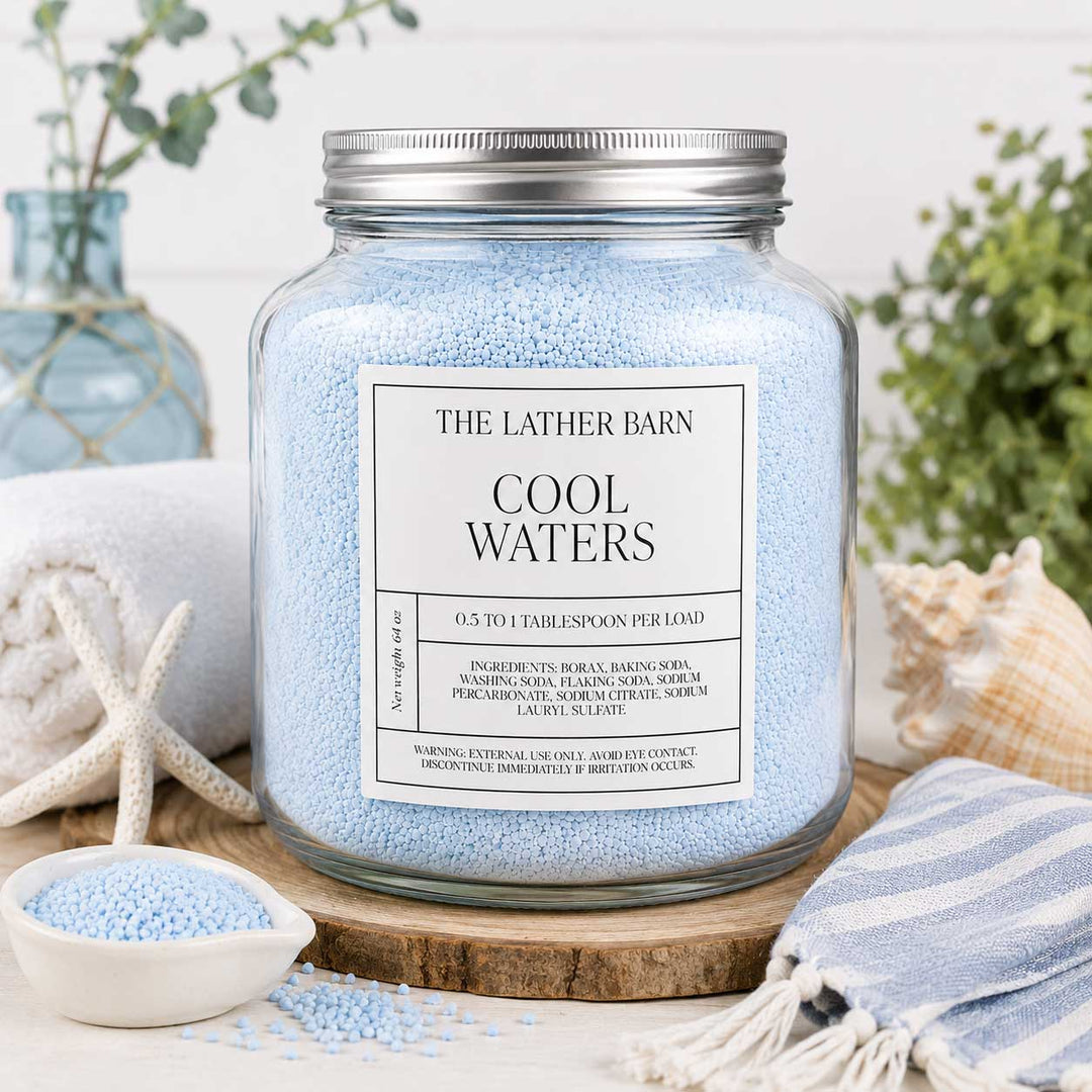 A large clear glass jar filled with light blue bath beads, labeled 'THE LATHER BARN COOL WATERS,' styled on a wooden board with a striped towel, starfish, seashells, and a small dish of blue beads.