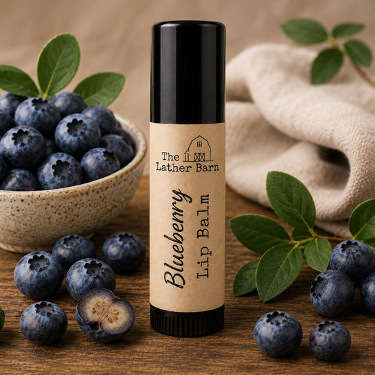 A tube of blueberry lip balm from The Lather Barn stands upright on a wooden surface, surrounded by fresh blueberries, green leaves, a bowl of blueberries, and a beige cloth in the background.