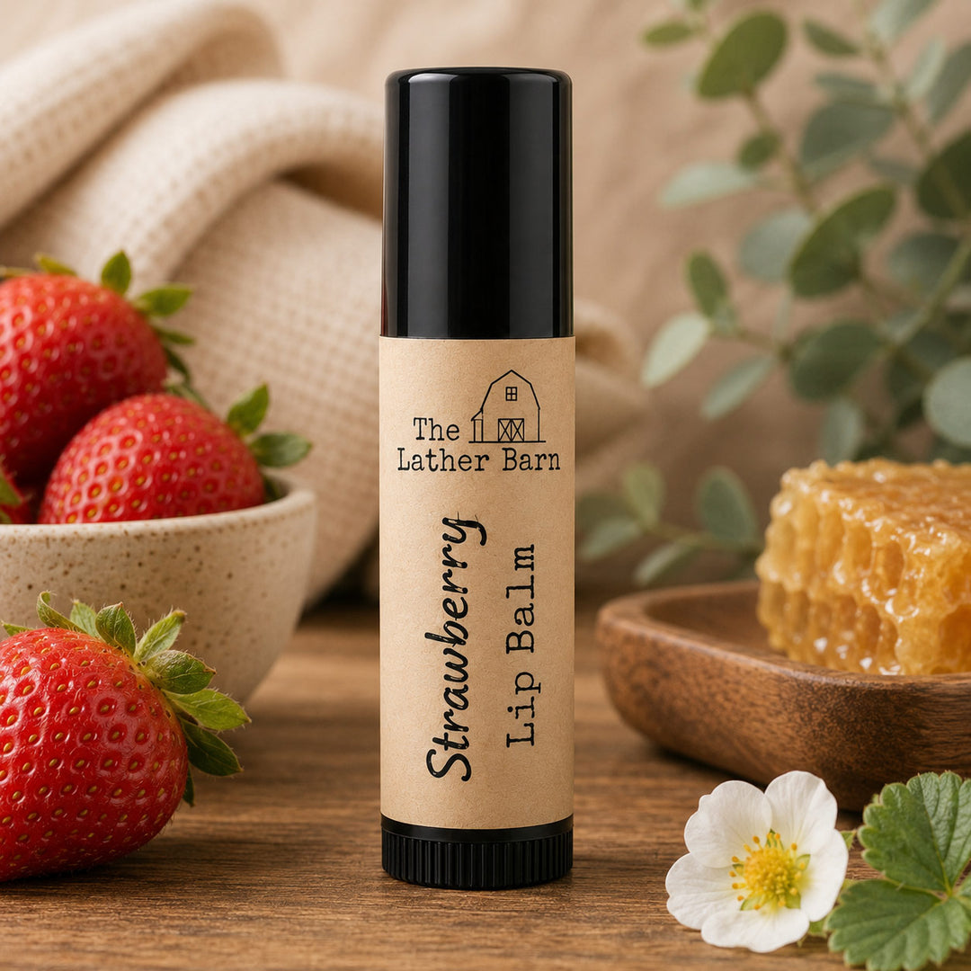 A tube of Strawberry Lip Balm by The Lather Barn stands on a wooden surface, surrounded by fresh strawberries, a bowl of honey, a white flower, and green leaves. The lip balm tube has a black cap and base with a beige label.