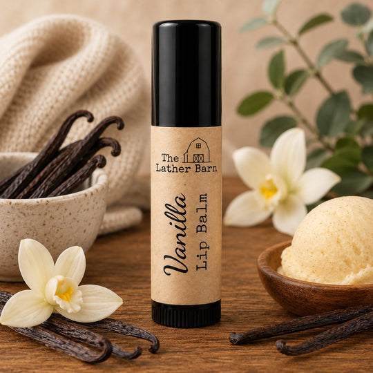 A tube of Vanilla Lip Balm by The Lather Barn is centered on a rustic wooden table, surrounded by vanilla beans, white flowers, a scoop of vanilla ice cream, and a beige knit fabric in the background.