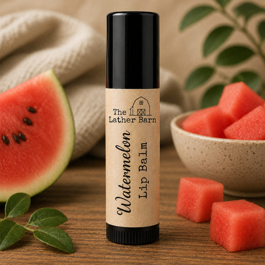 A tube of Watermelon Lip Balm by The Lather Barn stands on a wooden surface, surrounded by watermelon slices, a bowl of watermelon cubes, and green leaves in a styled setting.