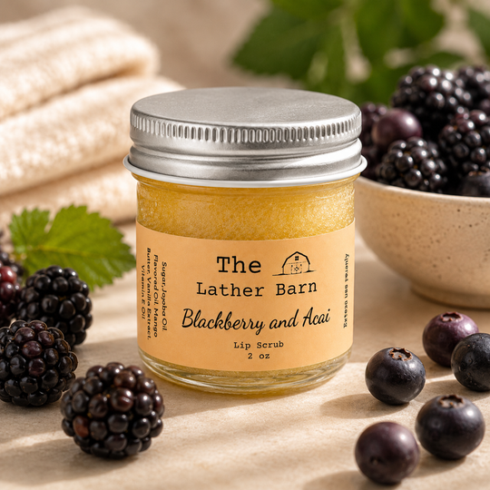 A jar of The Lather Barn Blackberry and Acai Lip Scrub sits on a beige surface surrounded by fresh blackberries, blueberries, and green leaves. The product is in a clear glass jar with a silver metal lid and a light orange label.
