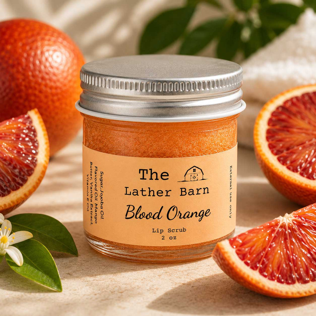 A jar of The Lather Barn Blood Orange lip scrub sits on a neutral surface surrounded by fresh blood orange slices, a whole orange, and green leaves. The product is in a clear glass jar with a silver metal lid and a beige label.