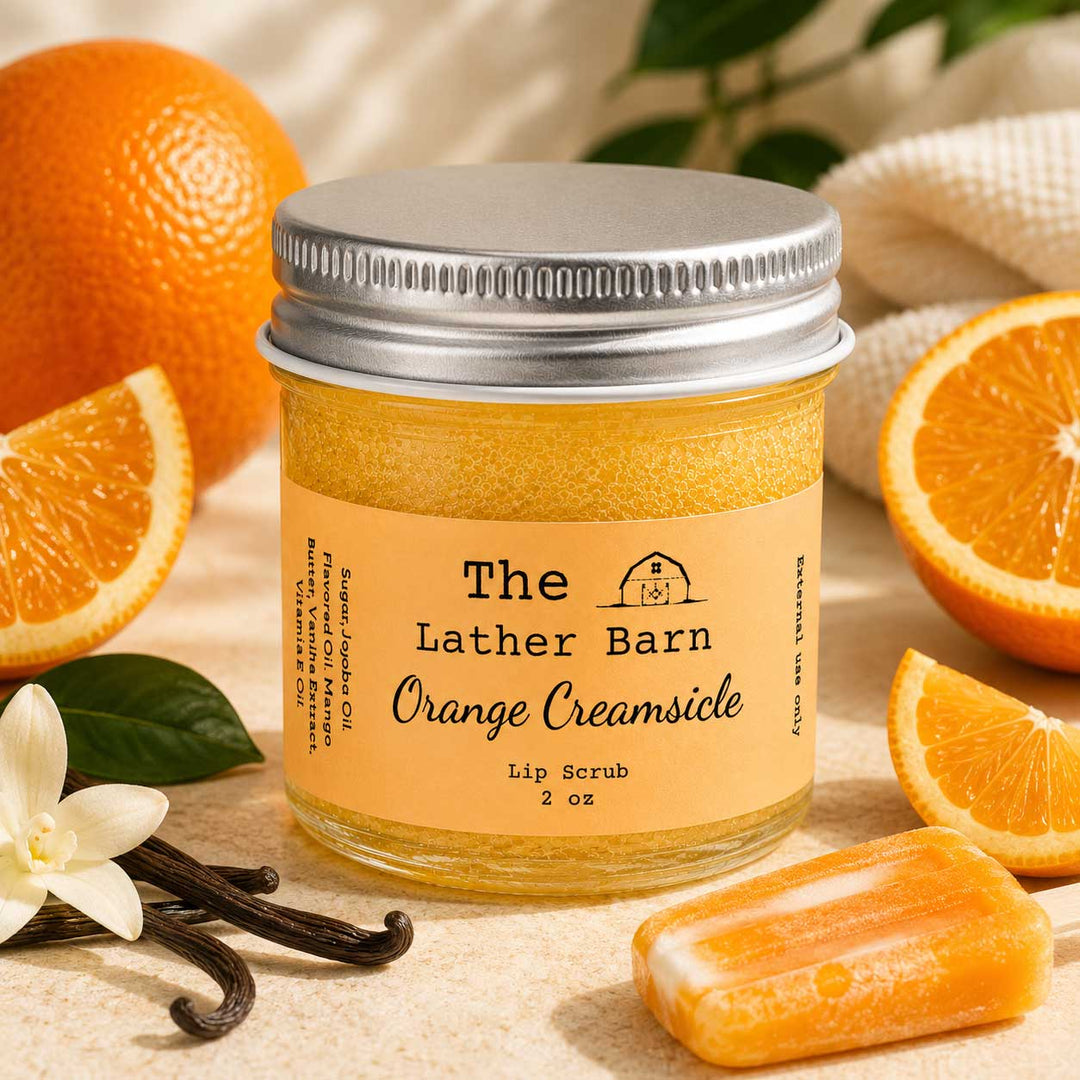A jar of The Lather Barn Orange Creamsicle lip scrub sits on a beige surface surrounded by orange slices, a white flower, vanilla beans, and a bar of orange soap. The jar has a silver metal lid and a light orange label with black text.