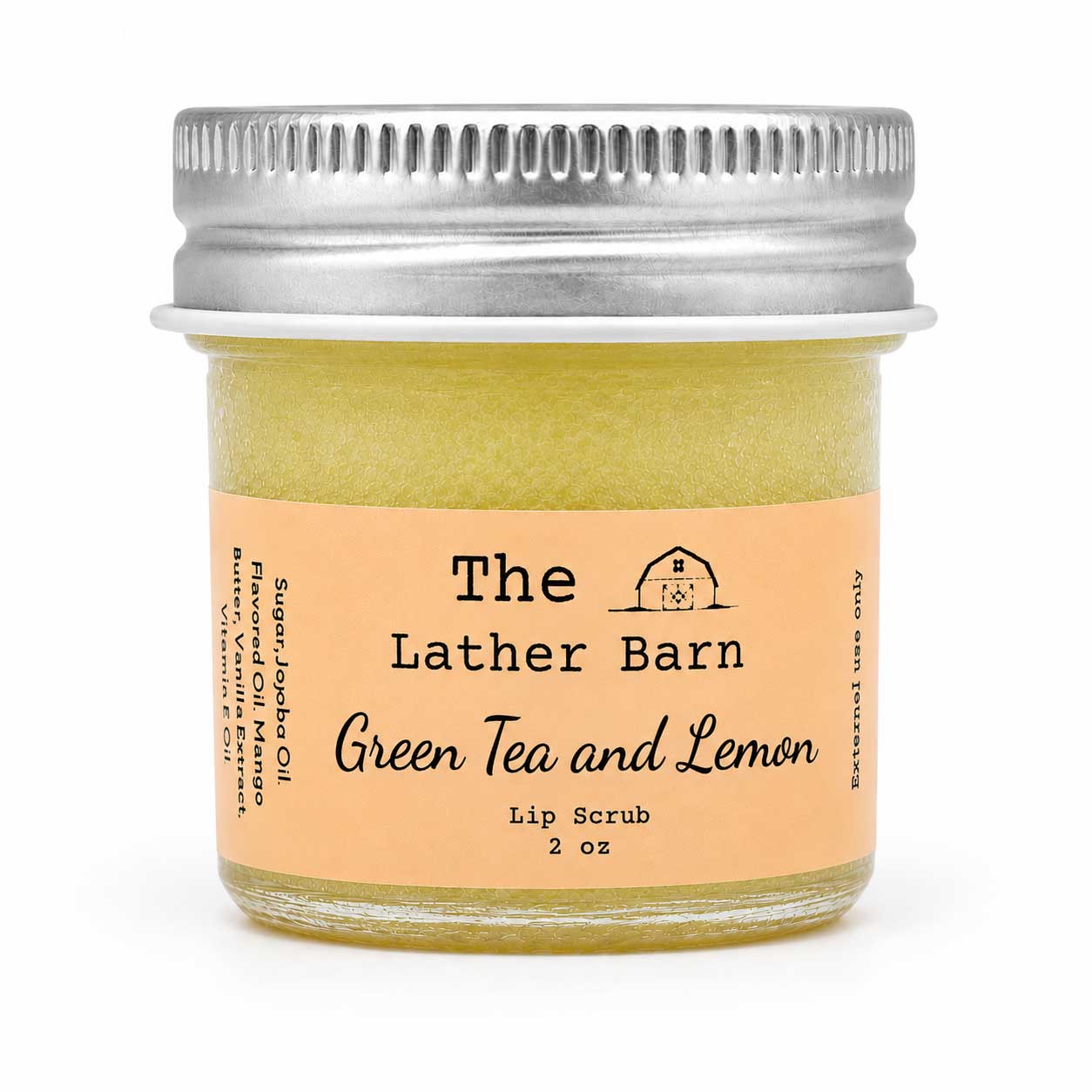 A small glass jar of lip scrub with a silver metal lid and a peach label reading 'The Lather Barn Green Tea and Lemon Lip Scrub 2 oz' on a white background.