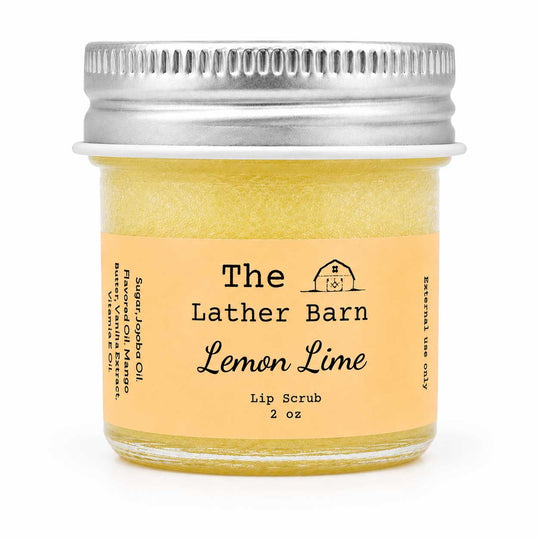 A 2 oz glass jar of The Lather Barn Lemon Lime Lip Scrub, filled with a pale yellow-green scrub and featuring a light peach label with a silver metal screw lid, set against a plain white background.