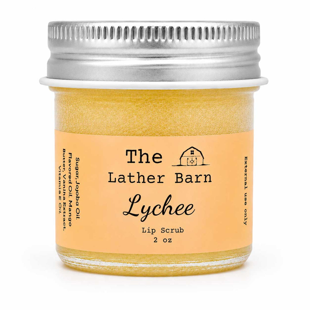 A 2 oz glass jar of The Lather Barn Lychee Lip Scrub, filled with a golden-yellow scrub and featuring a peach-colored label with a silver metal screw cap, set against a plain white background.
