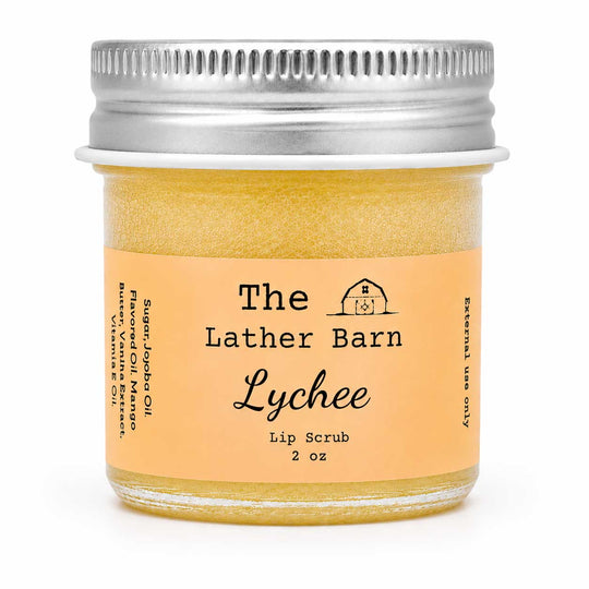 A 2 oz glass jar of The Lather Barn Lychee Lip Scrub, filled with a golden-yellow scrub and featuring a peach-colored label with a silver metal screw cap, set against a plain white background.