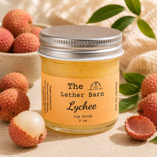 A jar of Lychee Lip Scrub by The Lather Barn sits on a neutral fabric surface surrounded by fresh lychees and green leaves. The product is in a clear glass jar with a silver metal lid and a warm peach-colored label.