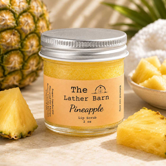 A jar of The Lather Barn Pineapple Lip Scrub sits on a light stone surface with pineapple slices and a pineapple rind around it. The product is in a clear glass jar with a silver metal lid and a peach-colored label.