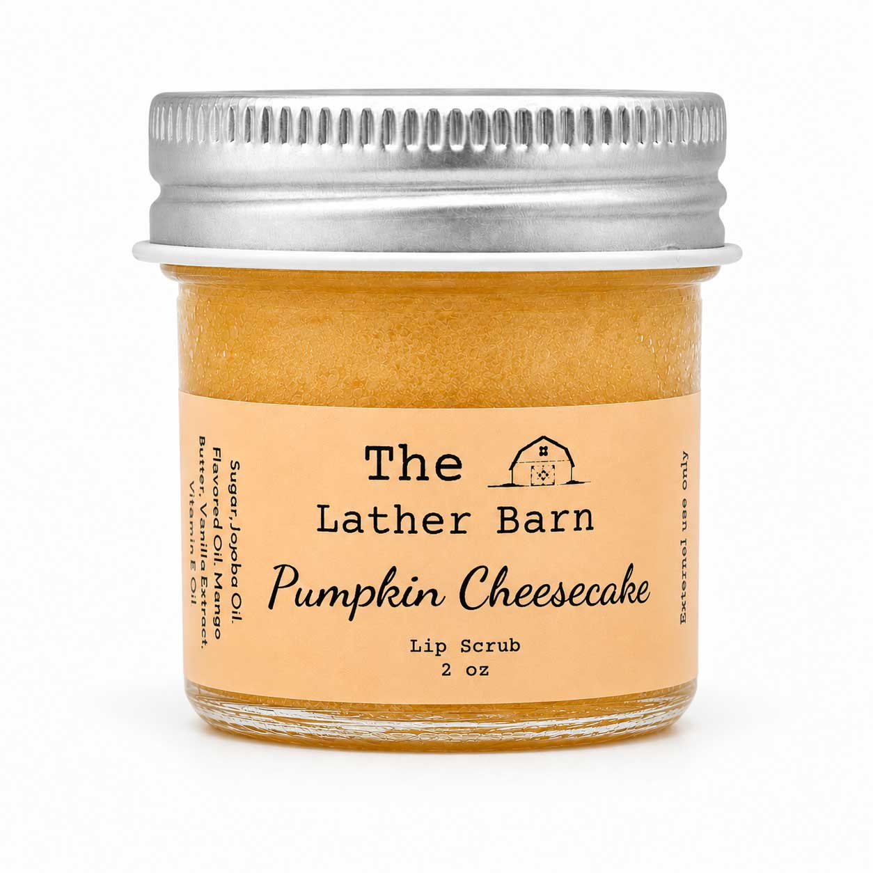 A 2 oz glass jar of The Lather Barn Pumpkin Cheesecake Lip Scrub, featuring a light peach label and a silver metal screw-on lid, set against a plain white background.