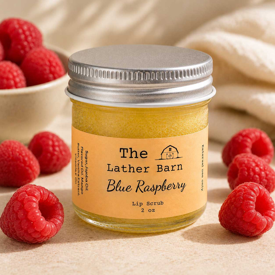 A jar of The Lather Barn Blue Raspberry lip scrub sits on a neutral surface surrounded by fresh raspberries, with a soft cream fabric blurred in the background.
