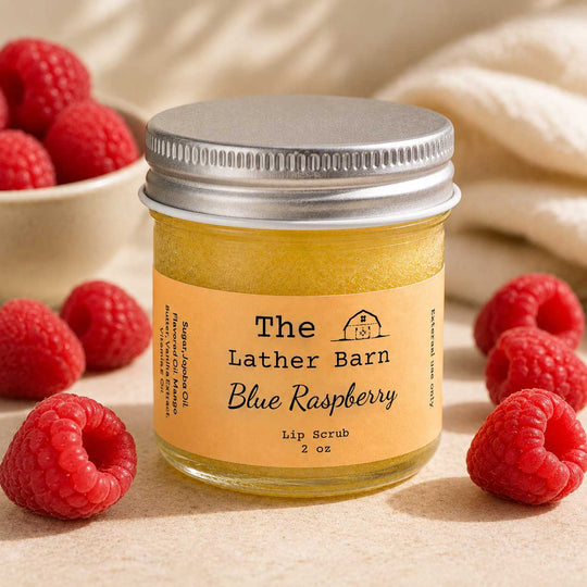 A jar of The Lather Barn Blue Raspberry lip scrub sits on a neutral surface surrounded by fresh raspberries, with a soft cream fabric blurred in the background.