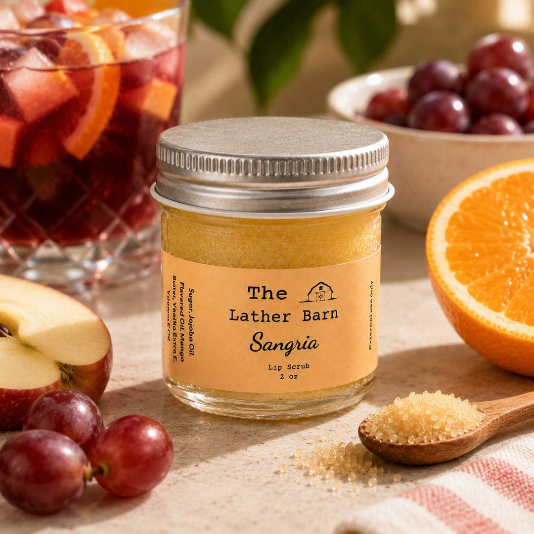 A jar of The Lather Barn Sangria lip scrub sits on a table surrounded by ingredients that make up the scent profile: sliced orange, red grapes, and an apple. A glass of sangria and a spoonful of brown sugar are also visible in the styled setting.