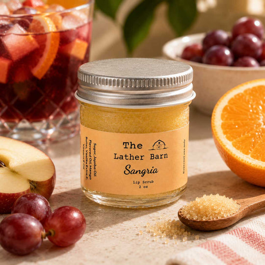 A jar of The Lather Barn Sangria lip scrub sits on a table surrounded by ingredients that make up the scent profile: sliced orange, red grapes, and an apple. A glass of sangria and a spoonful of brown sugar are also visible in the styled setting.