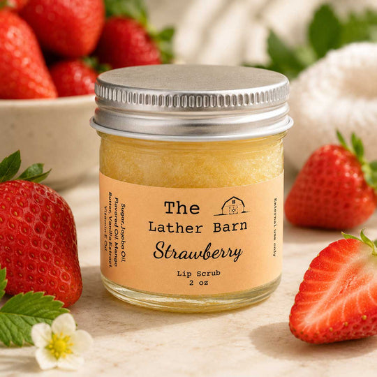 A small glass jar of strawberry lip scrub by The Lather Barn, surrounded by fresh strawberries on a light neutral surface. The jar has a silver metal lid and a tan label with black text.