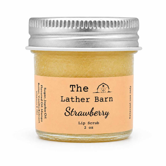 A small glass jar of strawberry lip scrub by The Lather Barn, filled with a pale yellowish-amber scrub and topped with a silver metal screw lid. The front label is peach-colored with black text.