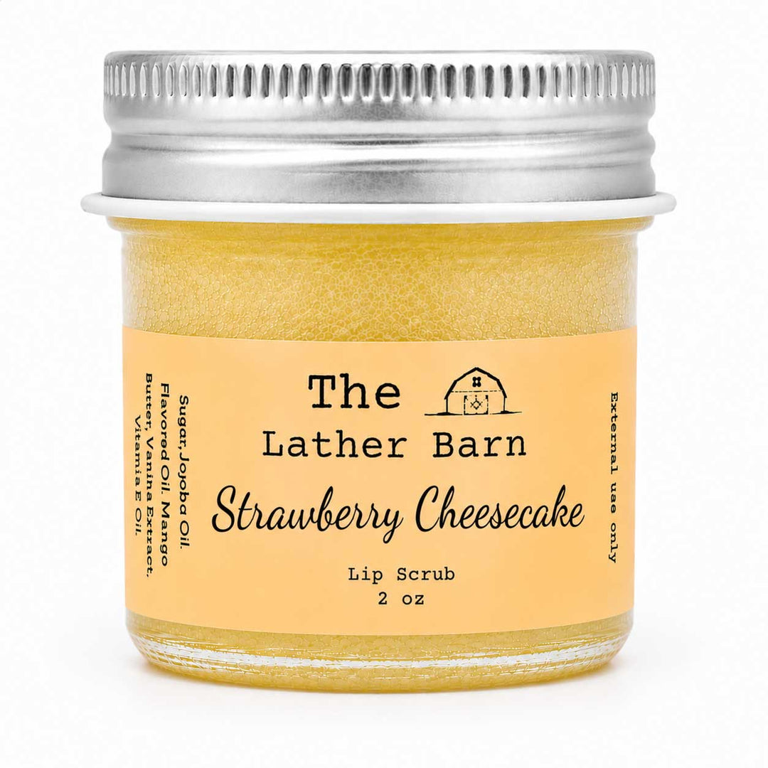 A 2 oz glass jar of The Lather Barn Strawberry Cheesecake lip scrub, filled with a yellowish-amber scrub and featuring a light peach label with black text and a silver metal screw cap, set against a plain white background.
