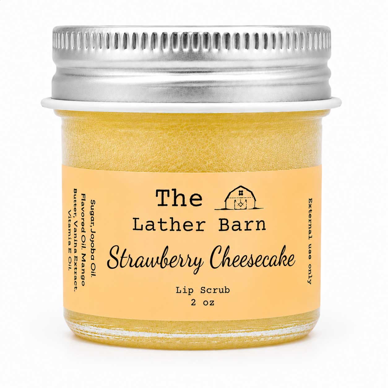 A 2 oz glass jar of The Lather Barn Strawberry Cheesecake lip scrub, filled with a yellowish-amber scrub and featuring a light peach label with black text and a silver metal screw cap, set against a plain white background.