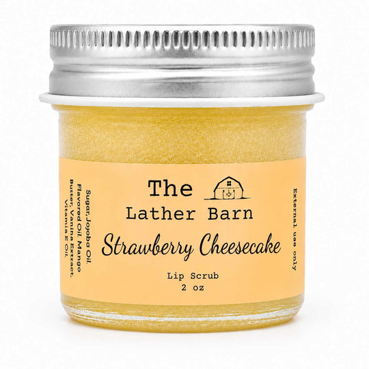 A 2 oz glass jar of The Lather Barn Strawberry Cheesecake lip scrub, filled with a yellowish-amber scrub and featuring a light peach label with black text and a silver metal screw cap, set against a plain white background.