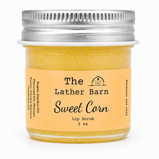 A 2 oz glass jar of The Lather Barn Sweet Corn Lip Scrub, filled with a golden yellow scrub and featuring a light peach label with a silver metal screw lid, set against a plain white background.