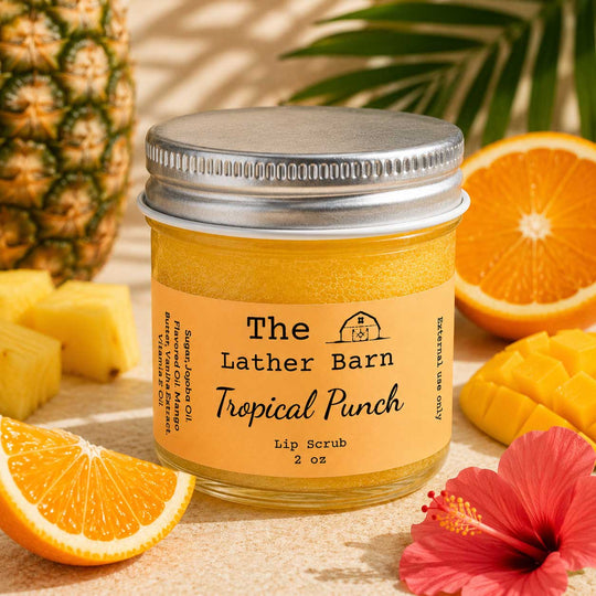 A jar of tropical punch lip scrub by The Lather Barn, surrounded by fresh pineapple, orange slices, mango pieces, and a red hibiscus flower on a sandy surface.