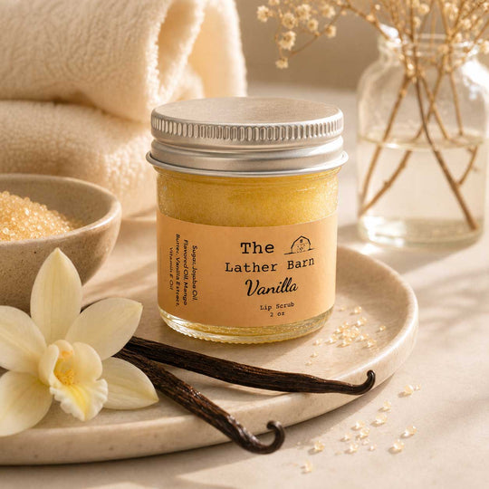 A jar of 'The Lather Barn Vanilla' lip scrub sits on a ceramic tray, styled with vanilla beans, a white orchid flower, and bath salts. The background features a soft beige towel and a glass vase with dried flowers.