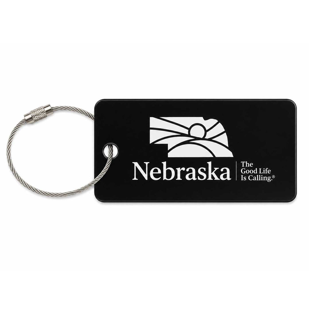 A black rectangular luggage tag with a silver metal cable loop, featuring the white Nebraska state outline, the word 'Nebraska', and the slogan 'The Good Life Is Calling'.