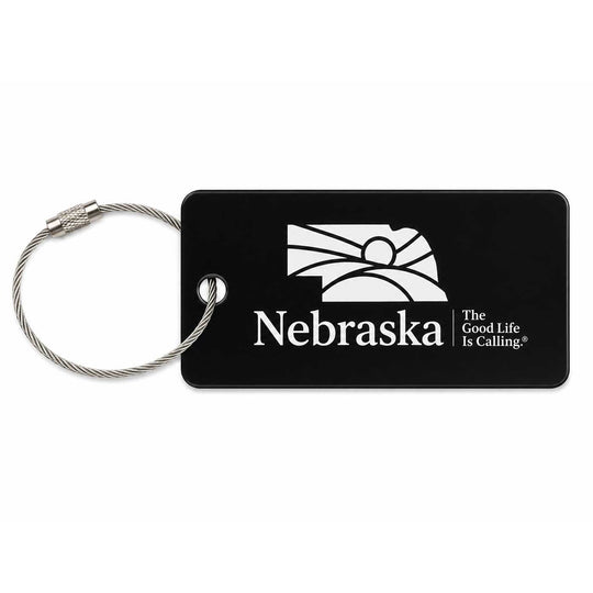 A black rectangular luggage tag with a silver metal cable loop, featuring the white Nebraska state outline, the word 'Nebraska', and the slogan 'The Good Life Is Calling'.