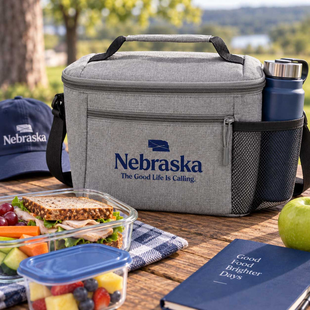 A gray insulated lunch bag with a top handle and shoulder strap sits on a wooden table outdoors. The bag has a front zip pocket with the Nebraska logo and text, and a side mesh pocket holding a dark blue water bottle.