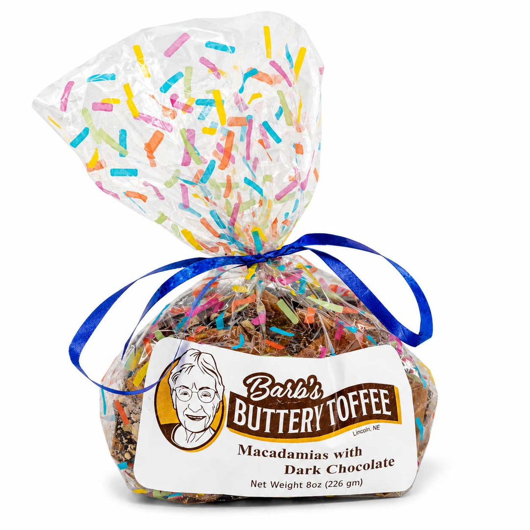 A packaged bag of Barb's Buttery Toffee Macadamias with Dark Chocolate, wrapped in a clear cellophane bag filled with colorful sprinkles and tied with a blue ribbon, on a white background.