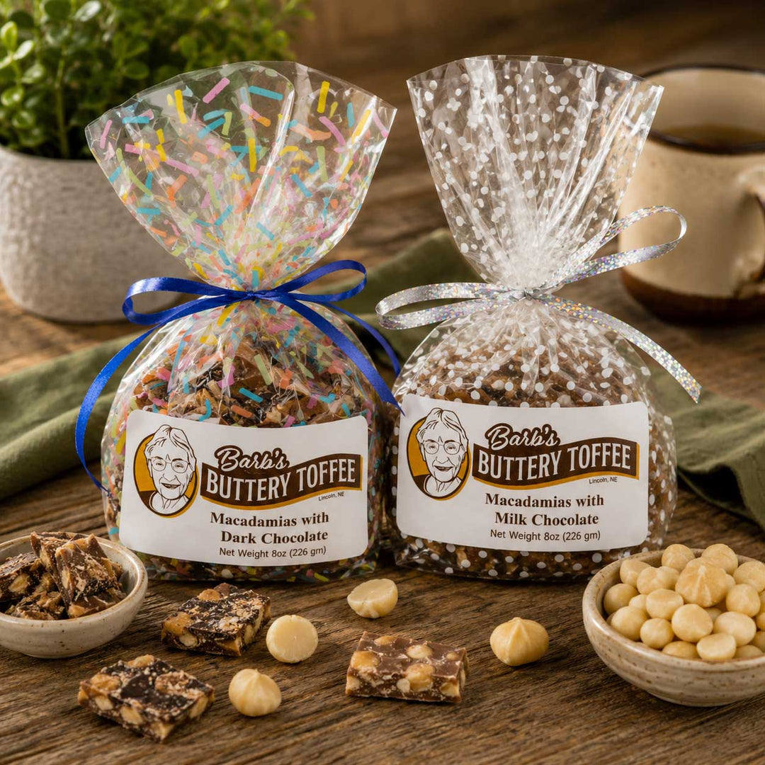 Two bags of Barb's Buttery Toffee are displayed on a rustic wooden table with bowls of ingredients and finished taffy squares. The bag on the left contains macadamias with dark chocolate and is tied with a blue ribbon, while the bag on the right contains macadamias with milk chocolate and is tied with a silver ribbon.