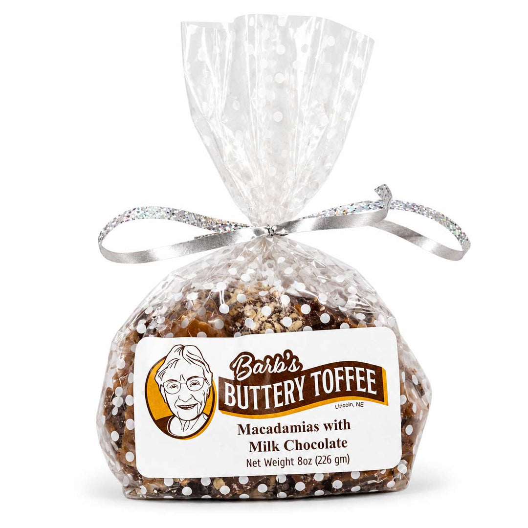 A 8 oz bag of Barb's Buttery Toffee Macadamias with Milk Chocolate, sealed in a clear cellophane bag tied with a silver glitter ribbon and featuring a white product label.