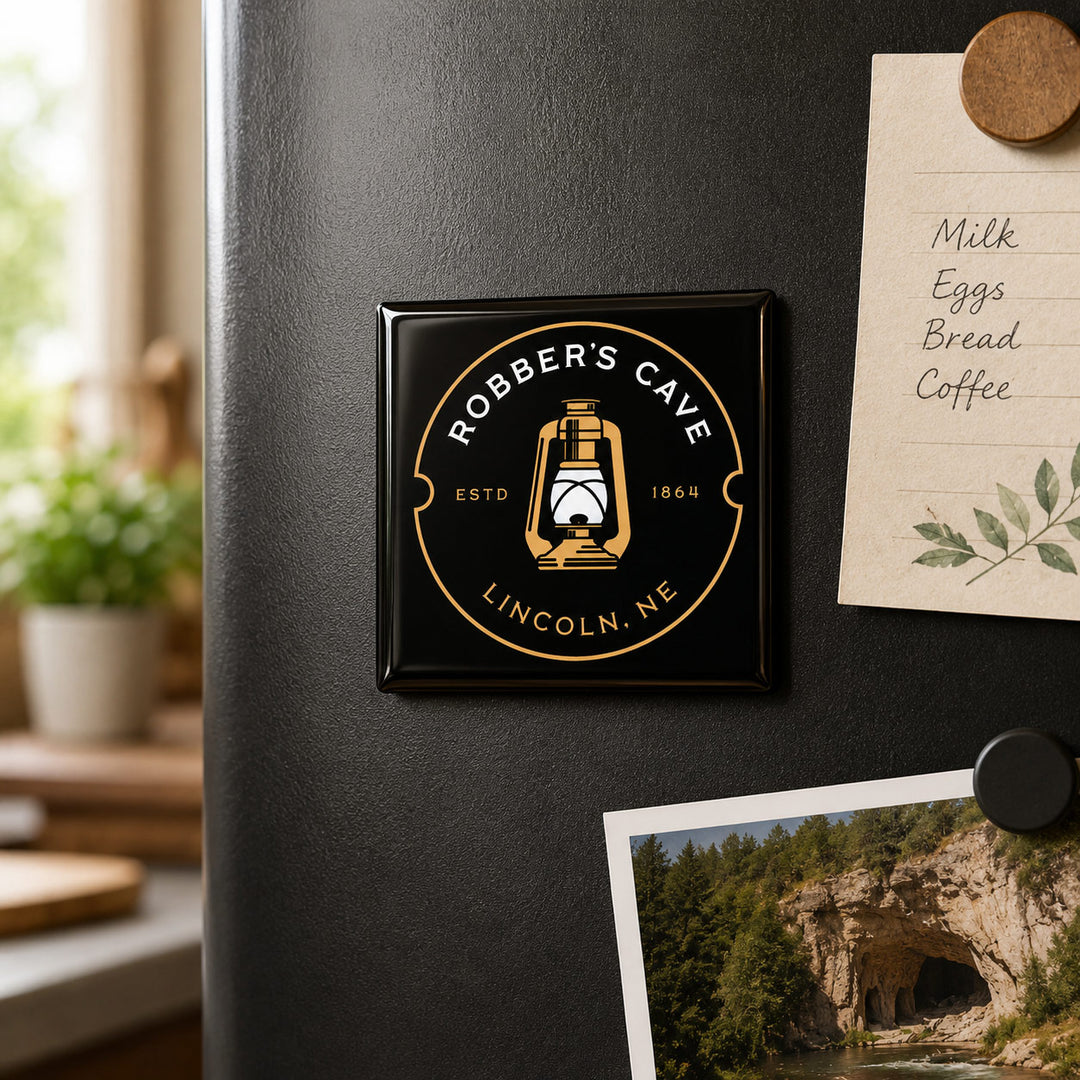 A square black refrigerator magnet featuring the 'Robber's Cave' logo in gold and white, attached to a dark gray refrigerator. The scene is styled as a cozy kitchen with a paper recipe card and a landscape photo pinned nearby.