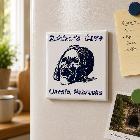 A square white refrigerator magnet with a dark blue skull illustration and the text 'Robber's Cave' and 'Lincoln, Nebraska', shown attached to a white refrigerator alongside other magnets and a sticky note.